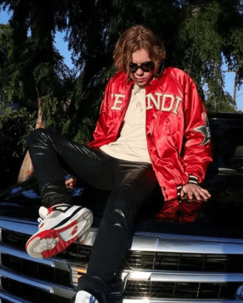 The Kid Laroi wearing a red satin varsity jacket sitting atop the hood of a car